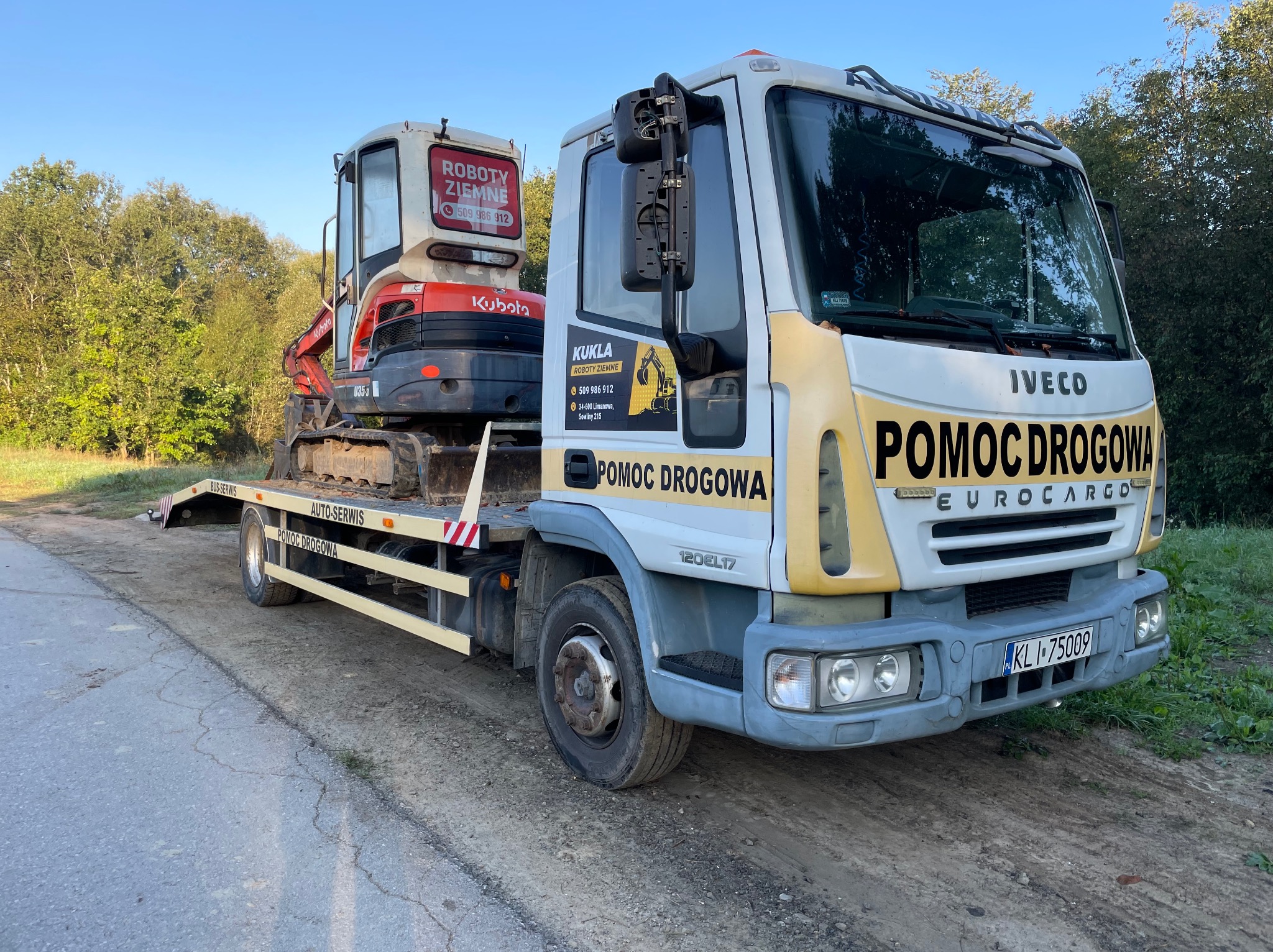 Ciężarówka pomocy drogowej marki Iveco z małą koparką Kubota na platformie, zaparkowana na poboczu drogi.