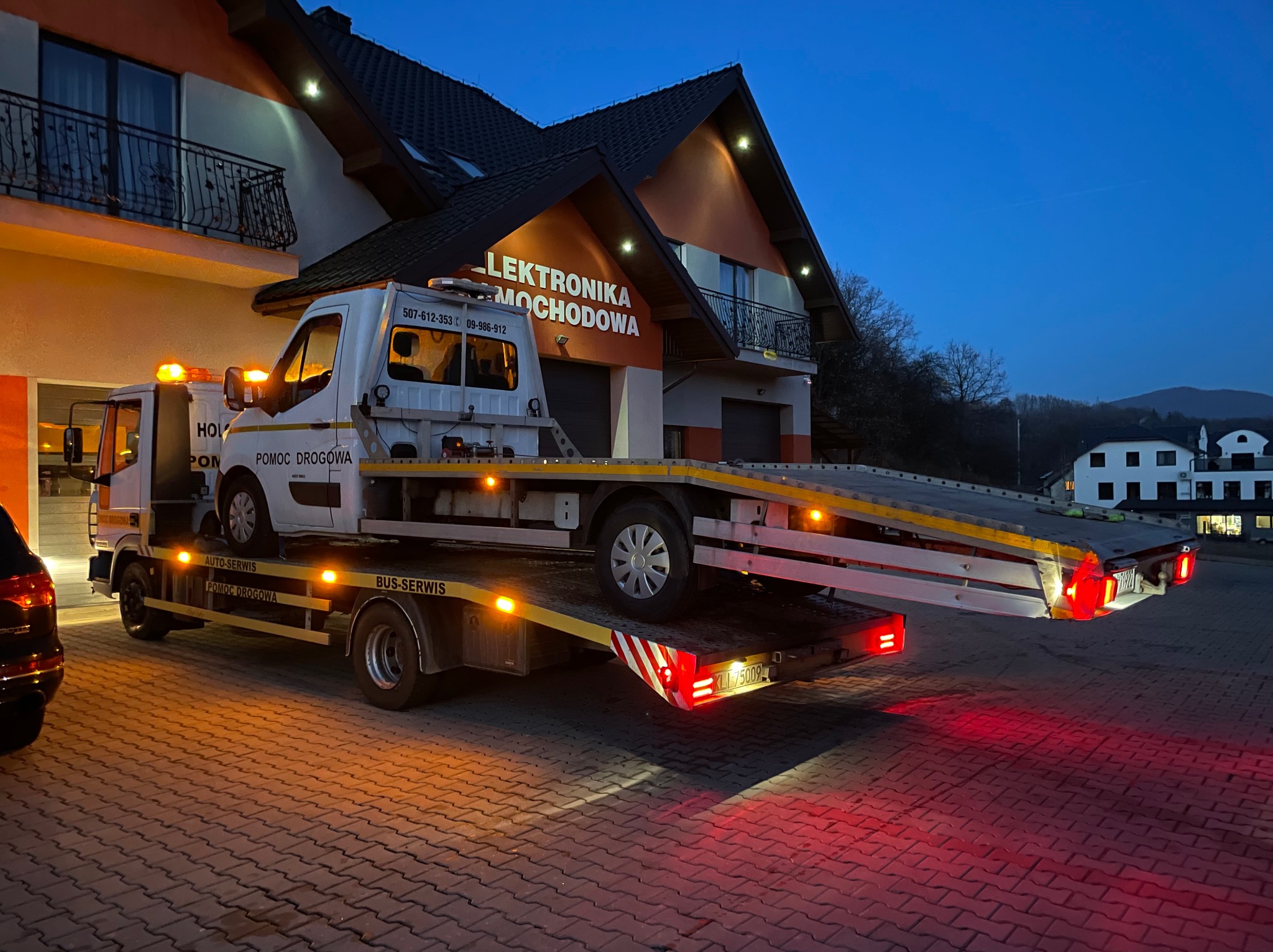 Biała laweta z pomarańczowymi światłami ostrzegawczymi przewozi uszkodzony samochód na tle budynku z napisem 'Elektronika Samochodowa' o zmierzchu.