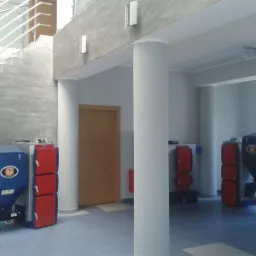 Wnętrze hali z kotłami na paliwo stałe, widoczne schody z balustradą nierdzewną i kolumny podtrzymujące strop.