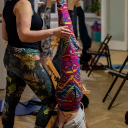 Instruktorka pomaga mężczyźnie w pozycji świecy, używając krzesła jako podpory, widoczne osoby w tle wykonujące to samo ćwiczenie