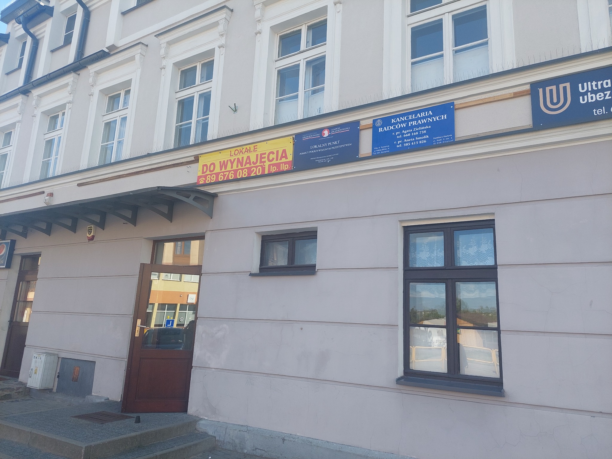 Fasada budynku z szyldami: Lokalny Punkt Pomocy Pokrzywdzonym Przestępstwem oraz Kancelaria Radców Prawnych z numerami telefonów i adresami, obok tablica 'Lokale do wynajęcia'.
