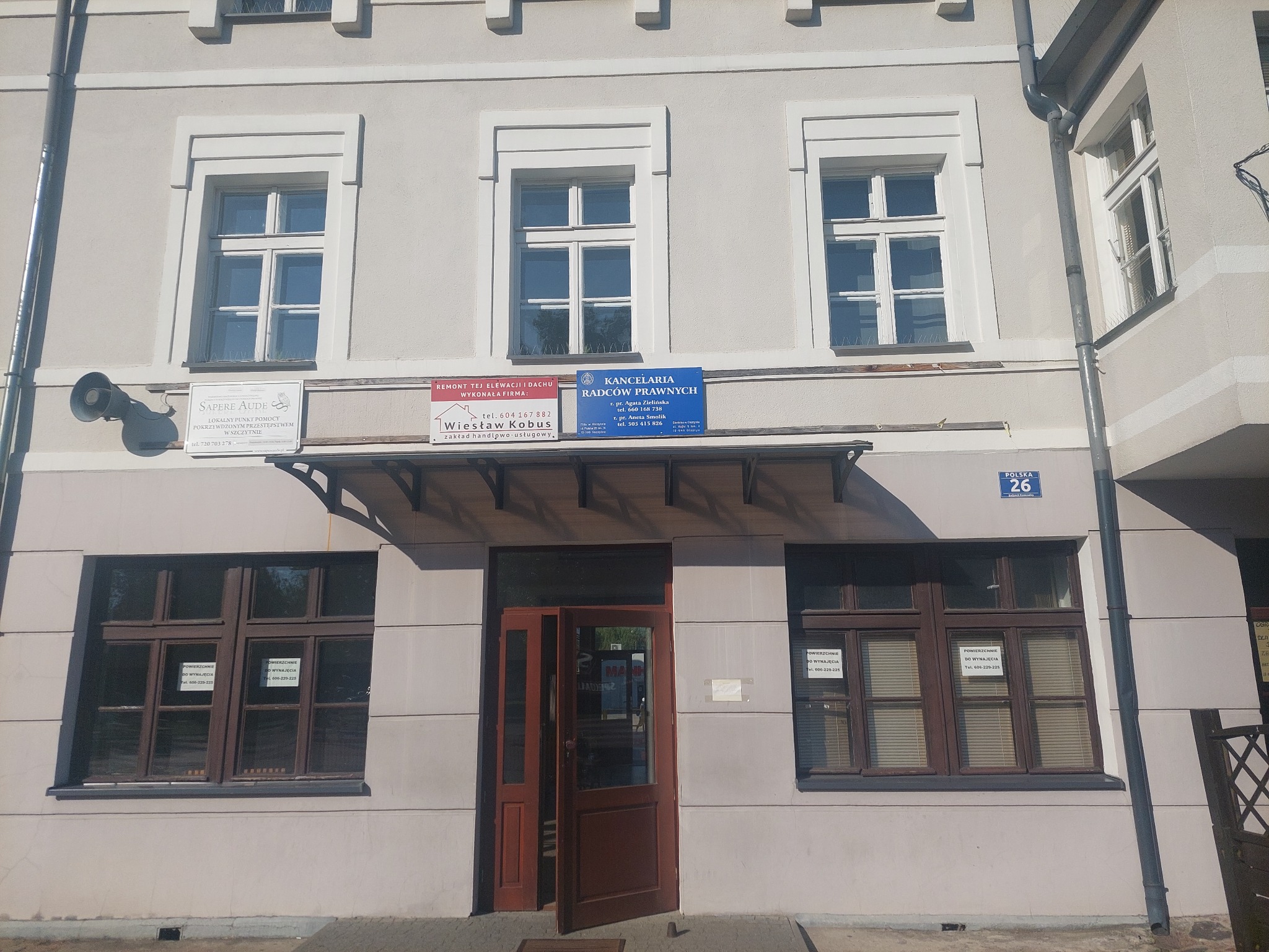 Fasada dwupiętrowego budynku z trzema szyldami kancelarii prawnych nad wejściem, okna z ogłoszeniami o wynajmie powierzchni.