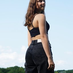 photo.recko - Młoda kobieta w czarnym topie i dżinsach, odwrócona tyłem, patrzy przez ramię na tle błękitnego nieba i zielonych drzew