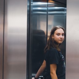 photo.recko - Portret młodej kobiety w czarnej koszulce i kwiecistej spódnicy, stojącej w nowoczesnej, metalowej windzie z torbą na ramieniu i delikatnym uśmiechem.