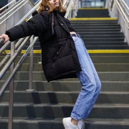 photo.recko - Młoda kobieta w zimowej kurtce i jeansach pozuje na schodach z metalową barierką, opierając się o nią.