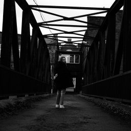 photo.recko - Czarno-białe zdjęcie kobiety w płaszczu stojącej na metalowym moście na tle budynków z cegły.