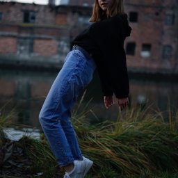 photo.recko - Młoda kobieta w czarnej bluzie i jeansach pozuje na tle zrujnowanego ceglanego budynku i zarośli, pochylona do przodu.