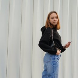 photo.recko - Młoda kobieta w czarnej bluzie z kapturem i jeansach pozuje z telefonem na tle białej, pionowo żebrowanej ściany.