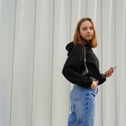 Młoda kobieta w czarnej bluzie z kapturem i jeansach pozuje z telefonem na tle białej, pionowo żebrowanej ściany.