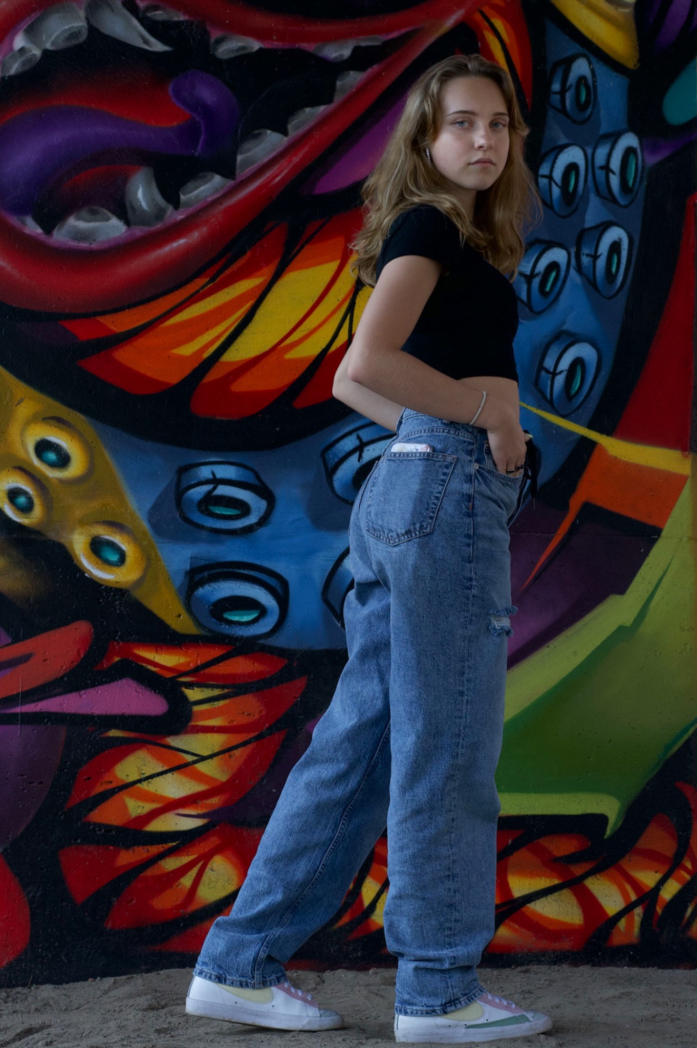 Młoda kobieta w jeansach i crop topie pozuje na tle barwnego graffiti z motywami oka i paszczy.