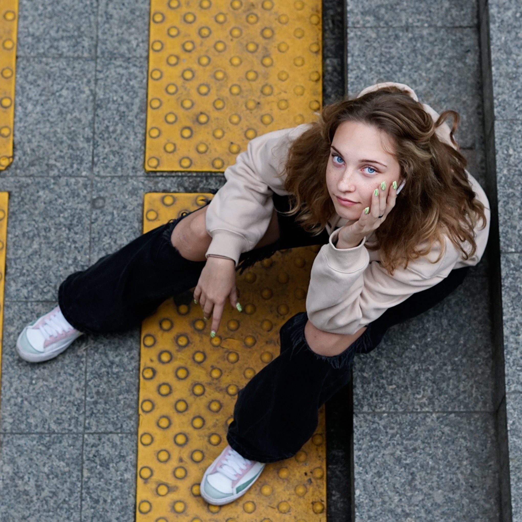 Portret młodej kobiety z błękitnymi oczami, siedzącej na chodniku z wypukłymi żółtymi elementami, ubrana w beżową bluzę i czarne spodnie z dziurami na kolanach, paznokcie pomalowane na zielono...