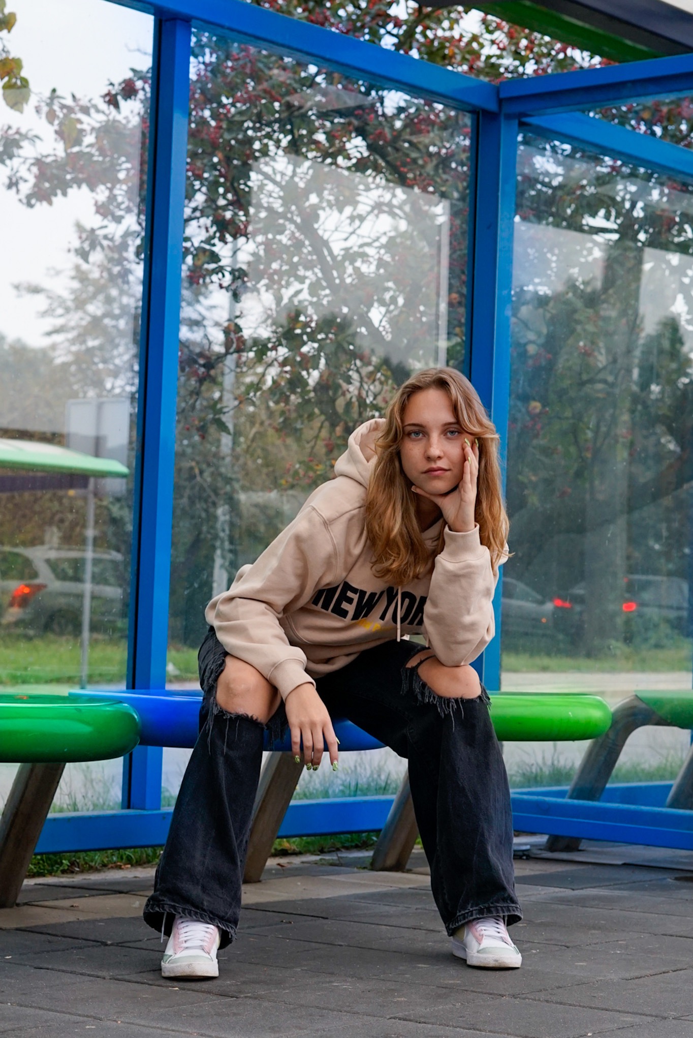 Młoda kobieta w bluzie z kapturem i podartych dżinsach siedzi na przystanku autobusowym, opierając brodę na dłoni i patrząc wprost w obiektyw.