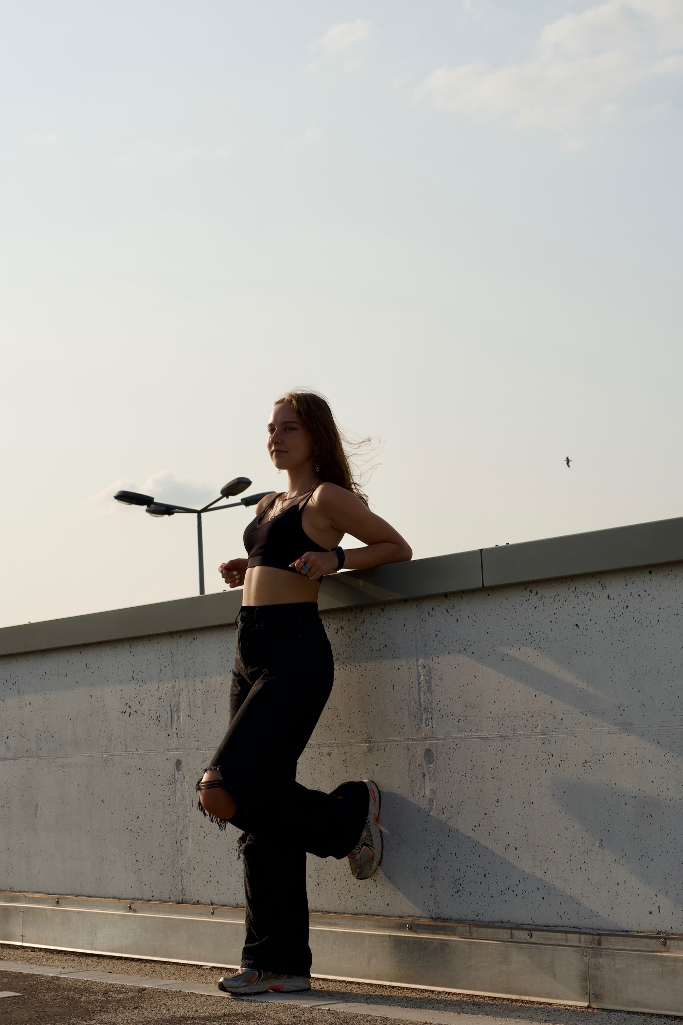 Młoda kobieta w czarnym stroju, oparta o betonową ścianę w Dobrej, z widocznymi lampami ulicznymi w tle.