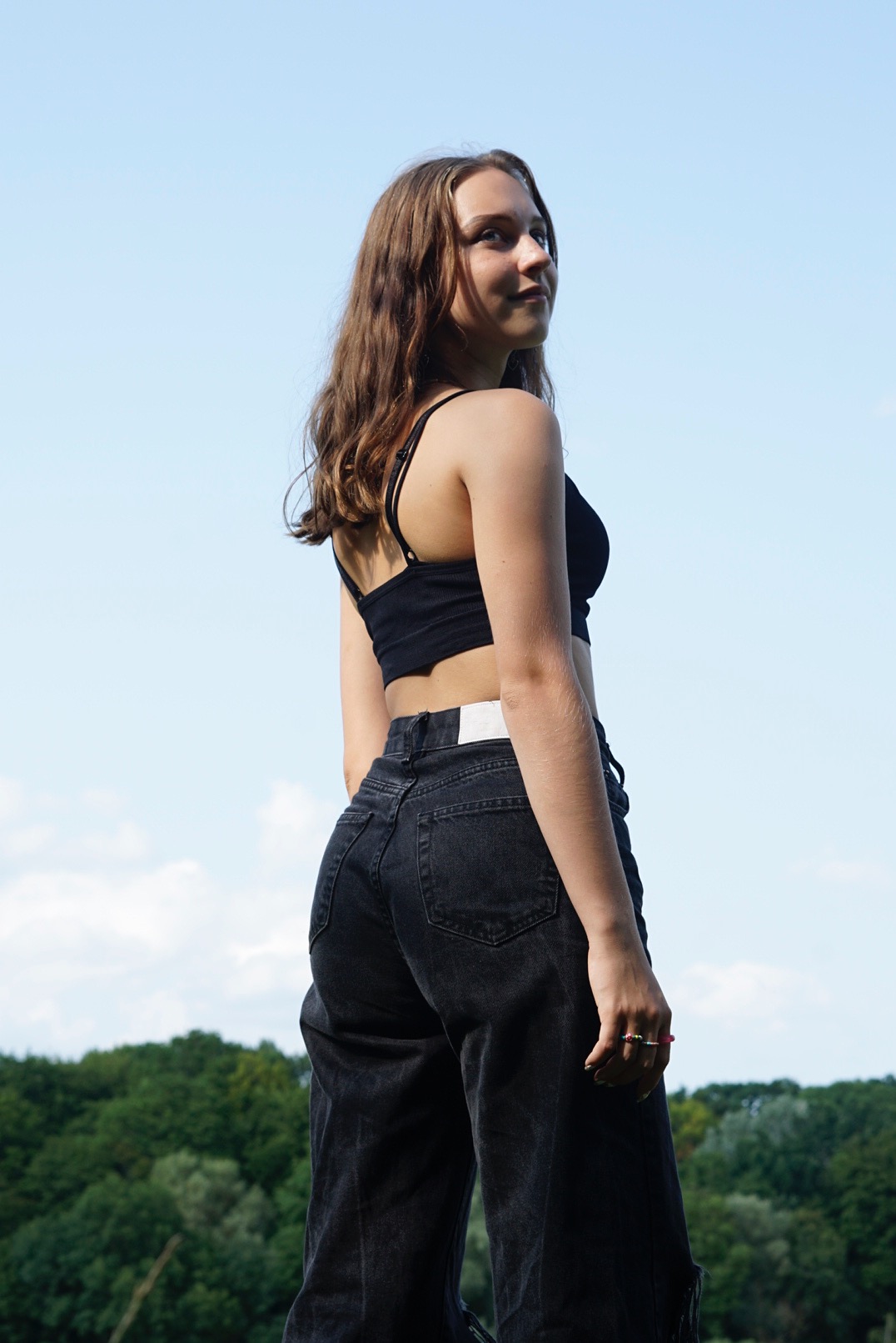 Młoda kobieta w czarnym topie i dżinsach, odwrócona tyłem, patrzy przez ramię na tle błękitnego nieba i zielonych drzew