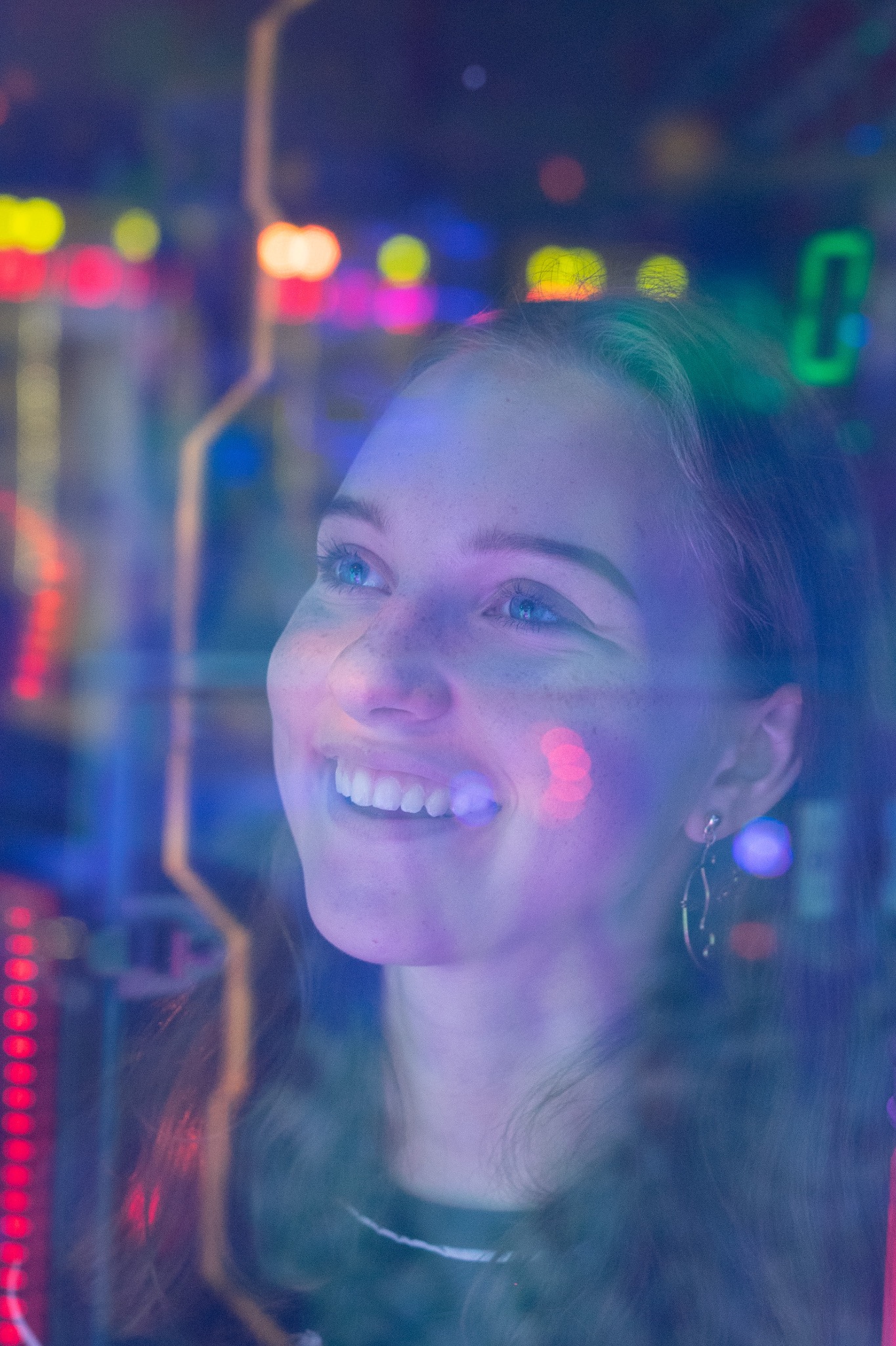 Portret uśmiechniętej kobiety z kolczykiem, sfotografowanej przez szybę z odbiciami kolorowych świateł i elementów automatu do gier.