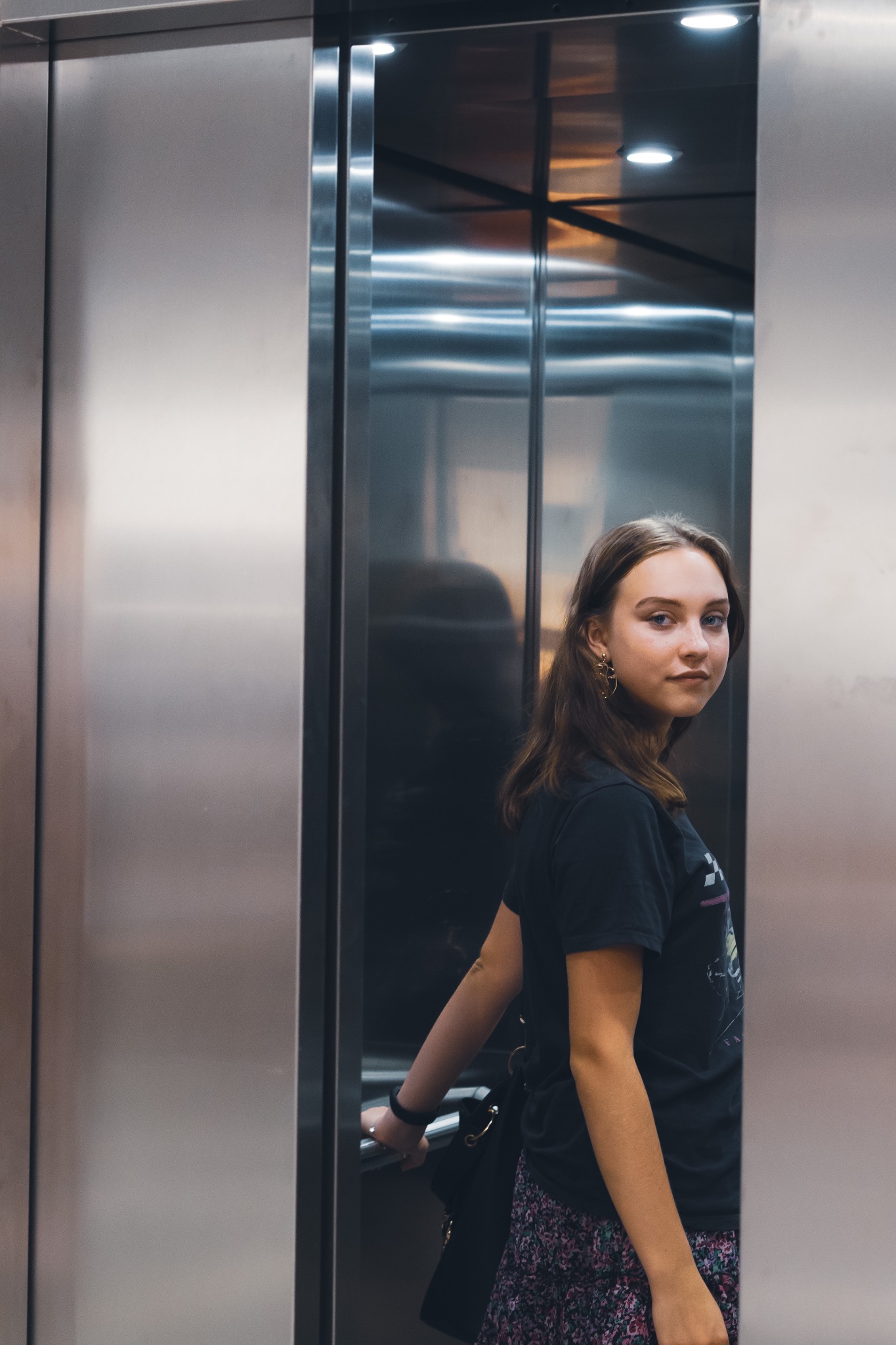 Portret młodej kobiety w czarnej koszulce i kwiecistej spódnicy, stojącej w nowoczesnej, metalowej windzie z torbą na ramieniu i delikatnym uśmiechem.