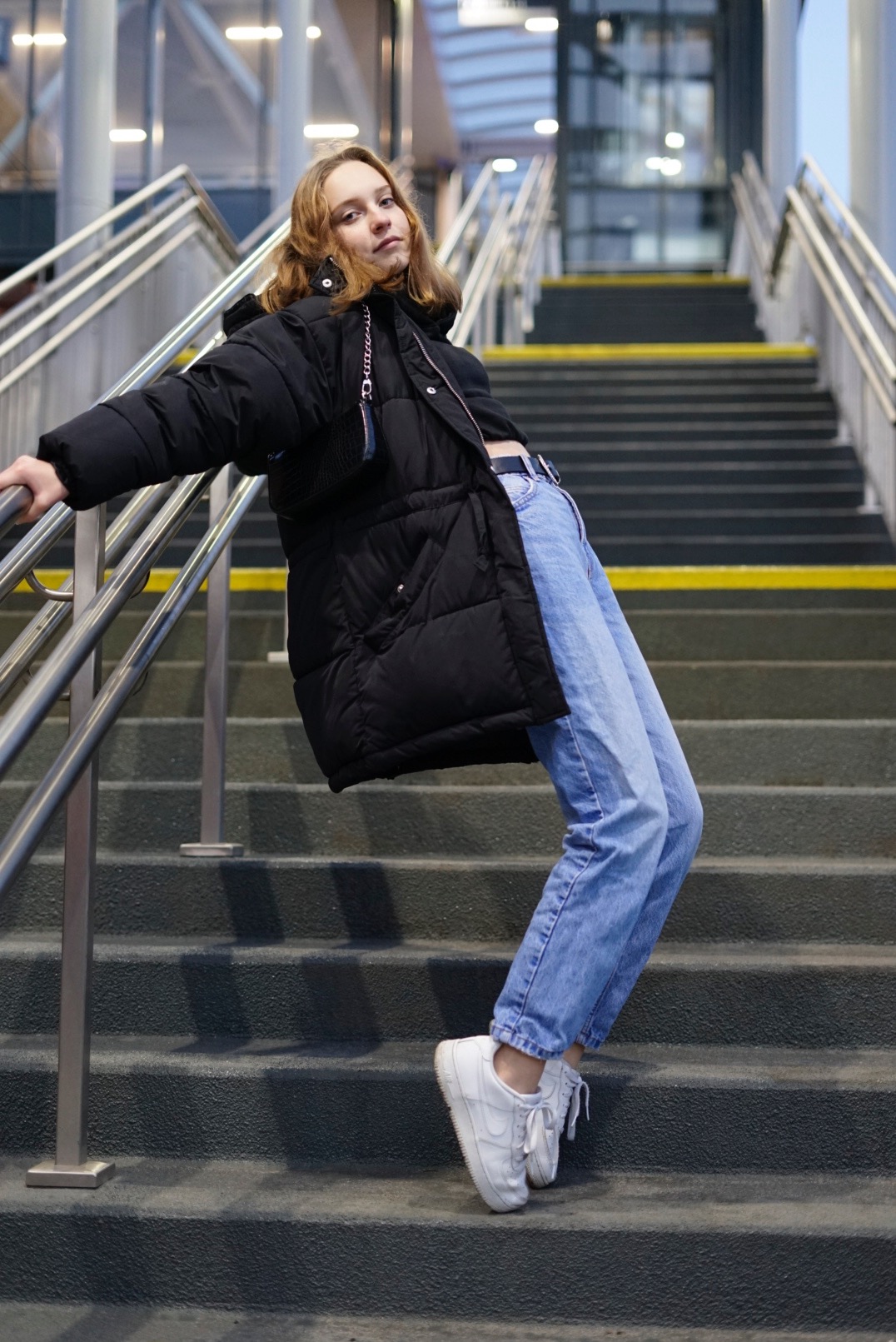 Młoda kobieta w zimowej kurtce i jeansach pozuje na schodach z metalową barierką, opierając się o nią.