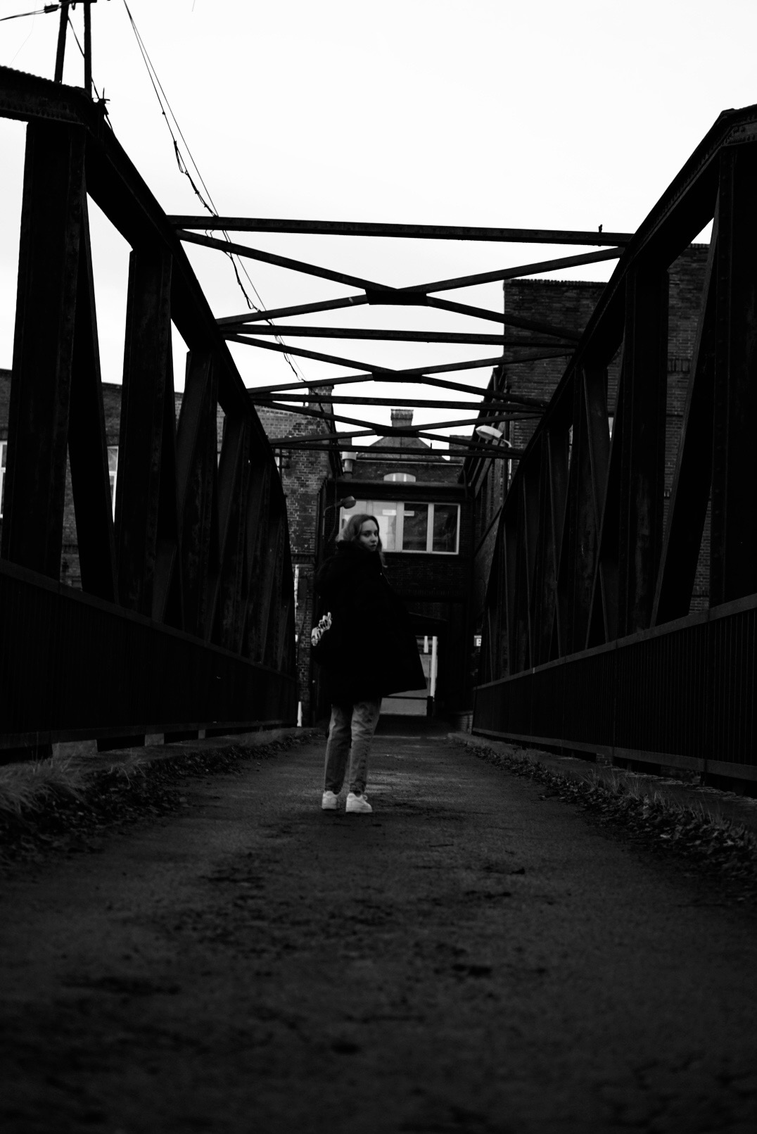 Czarno-białe zdjęcie kobiety w płaszczu stojącej na metalowym moście na tle budynków z cegły.