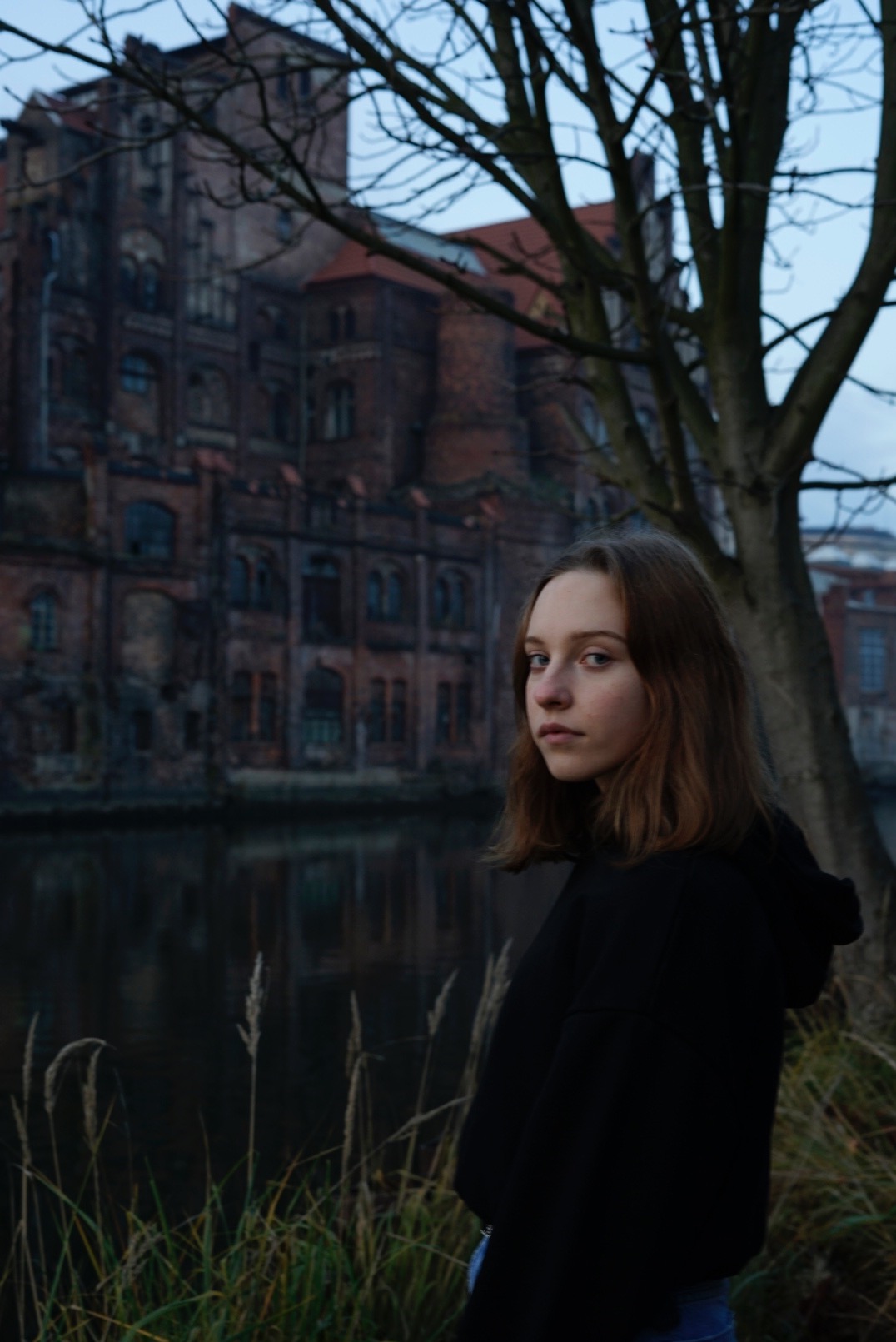 Portret młodej kobiety w czarnej bluzie z kapturem na tle starej, ceglanej fabryki i rzeki, pochmurny dzień.