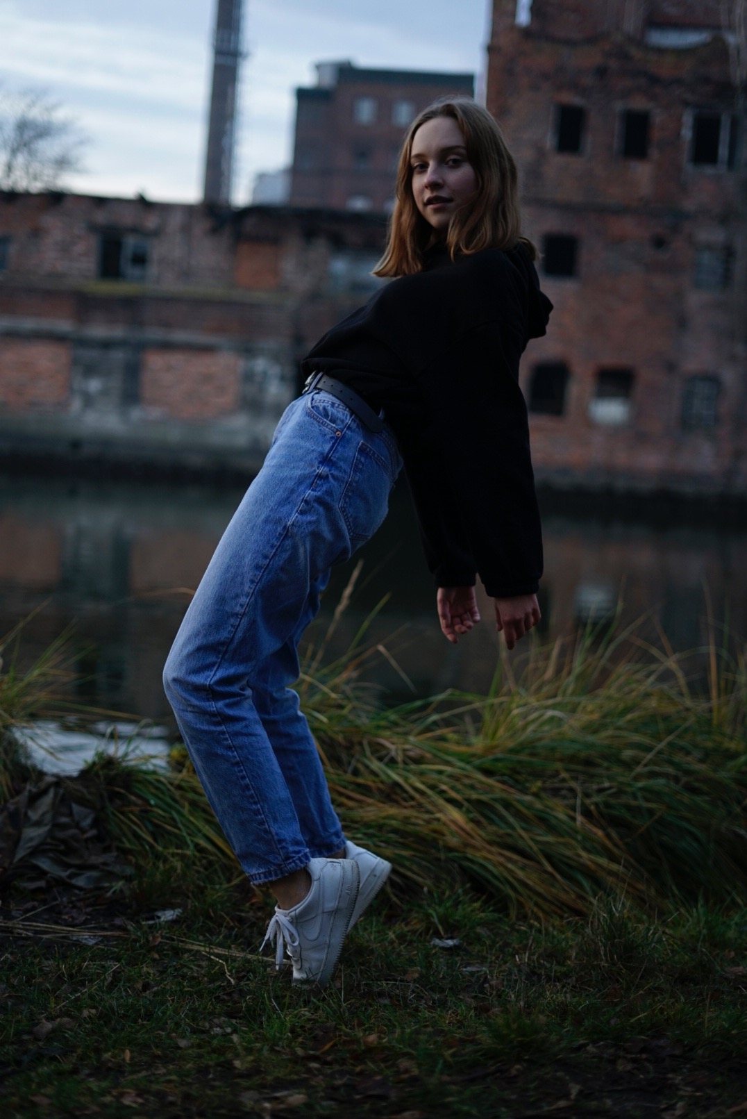 Młoda kobieta w czarnej bluzie i jeansach pozuje na tle zrujnowanego ceglanego budynku i zarośli, pochylona do przodu.