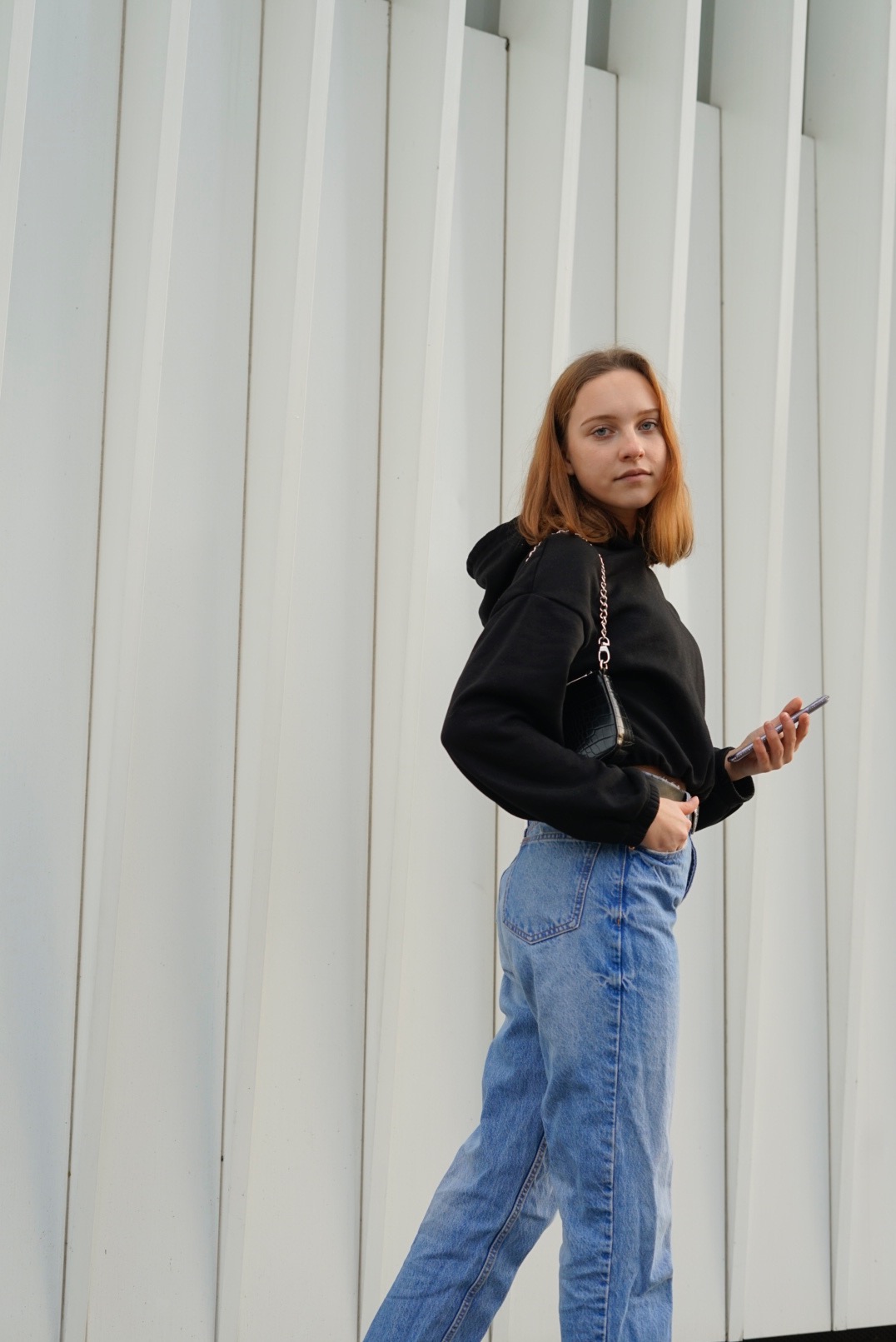 Młoda kobieta w czarnej bluzie z kapturem i jeansach pozuje z telefonem na tle białej, pionowo żebrowanej ściany.
