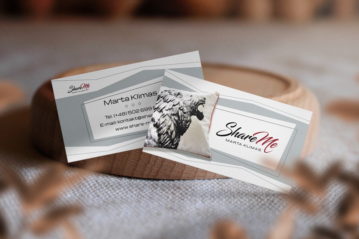 Dwie wizytówki z logo 'Share Me' i danymi kontaktowymi, ozdobione małą poduszką ze lwem, ułożone na drewnianej podstawce na tle tekstylnej powierzchni.