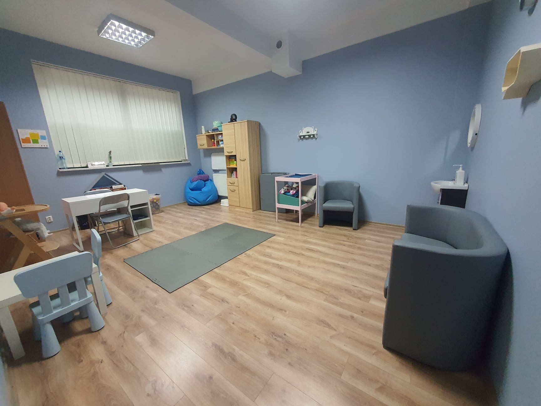Gabinet terapeutyczny z niebieskimi ścianami, drewnianą podłogą, wyposażony w stolik, krzesła dziecięce, pufę, szafę i dywanik piankowy
