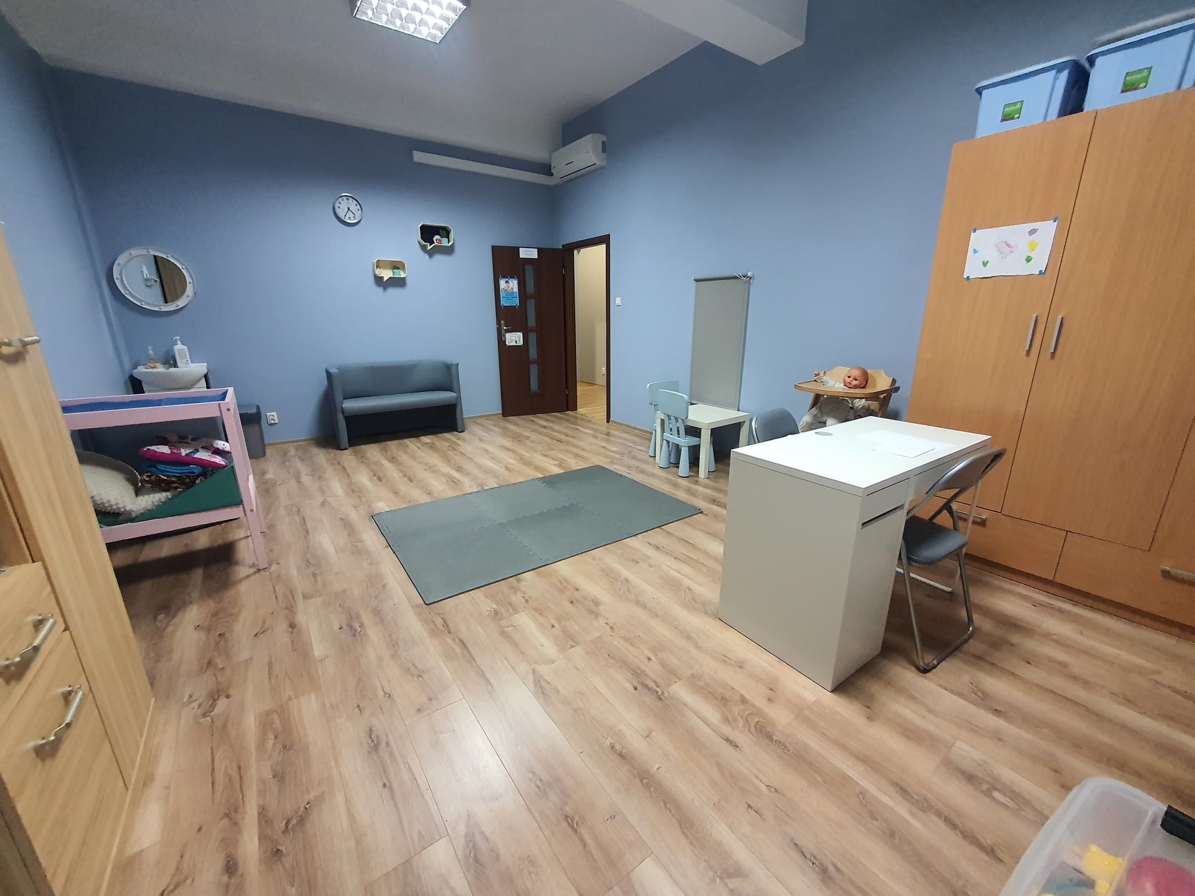 Przestronny gabinet z drewnianą podłogą, wyposażony w stolik dla dzieci, krzesła, sofę, szafę i dywan z puzzli, a także lalkę w krzesełku do karmienia.