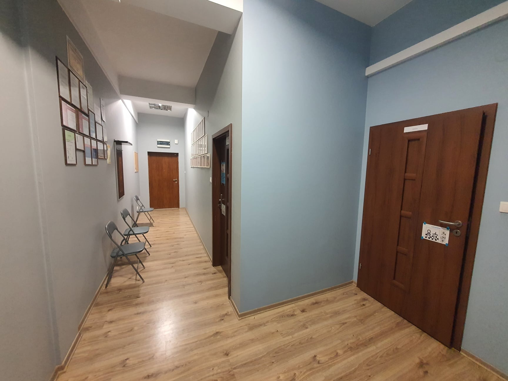 Korytarz w placówce medycznej z drewnianą podłogą, szarymi ścianami, niebieską ścianą, rzędem krzeseł i drzwiami z informacją o toalecie.