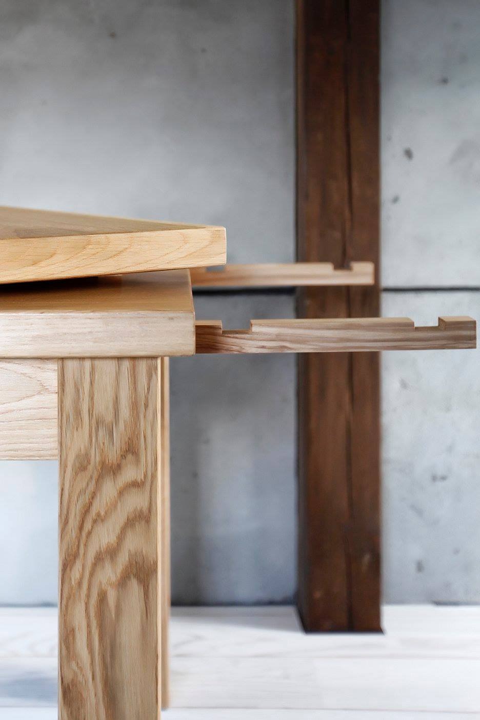 Fragment konstrukcji drewnianej z jasnego drewna, ukazujący elementy łączeniowe i strukturę słojów, na tle betonowej ściany i drewnianego filaru.