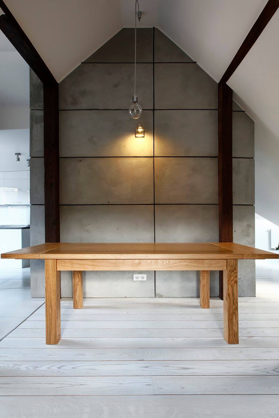 Jasny, dębowy stół jadalny z prostymi nogami, ustawiony na bielonej podłodze z desek, na tle ściany z betonowych płyt i wiszącej lampy z żarówką Edisona.