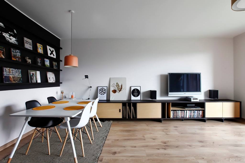 Nowoczesny salon z drewnianą podłogą, białym stołem jadalnym z kolorowymi podkładkami i szafką RTV z drewnianymi frontami oraz czarnymi regałami na książki.