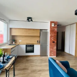 Nowoczesne wnętrze z aneksem kuchennym: biało-drewniane szafki, ceglana ściana, stół z nakryciem i niebieska sofa w tle.