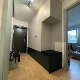 Wnętrze przedpokoju z zabudowaną szafą na wymiar w kolorach bieli i ciemnej zieleni, z siedziskiem i otwartymi półkami, na podłodze parkiet w jodełkę.