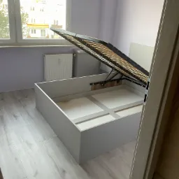 Otwarta rama łóżka z pojemnikiem, widoczny mechanizm podnoszenia stelaża, jasne panele podłogowe, fragment drzwi po prawej, okno w tle.