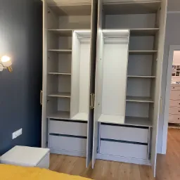 Dwie otwarte, jasnoszare szafy z półkami i szufladami, z oświetleniem LED wewnątrz, na tle ciemnej ściany i drewnianej podłogi, widoczny fragment żółtej narzuty na łóżko.