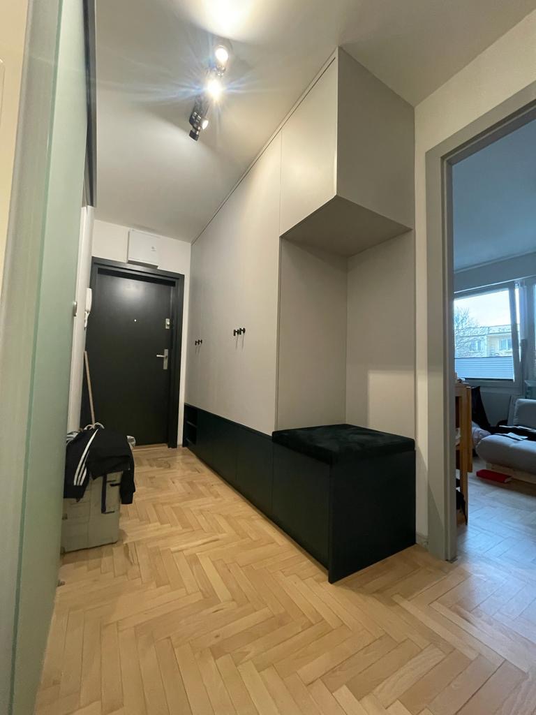 Wnętrze przedpokoju z zabudowaną szafą na wymiar w kolorach bieli i ciemnej zieleni, z siedziskiem i otwartymi półkami, na podłodze parkiet w jodełkę.