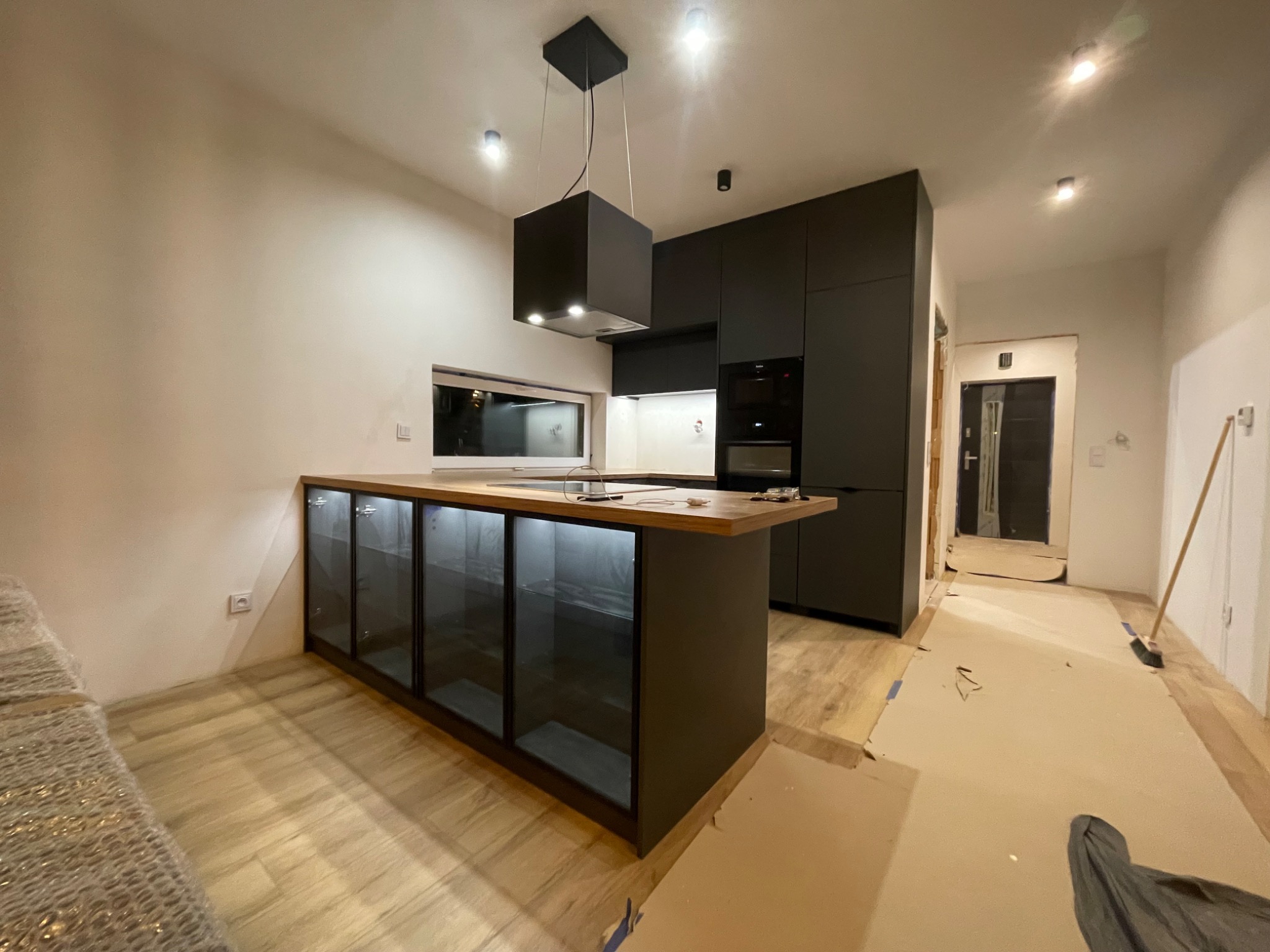 Nowoczesna kuchnia z czarnymi szafkami i wyspą z drewnianym blatem oraz szklanymi frontami, widoczna w trakcie montażu, podłoga zabezpieczona kartonem.