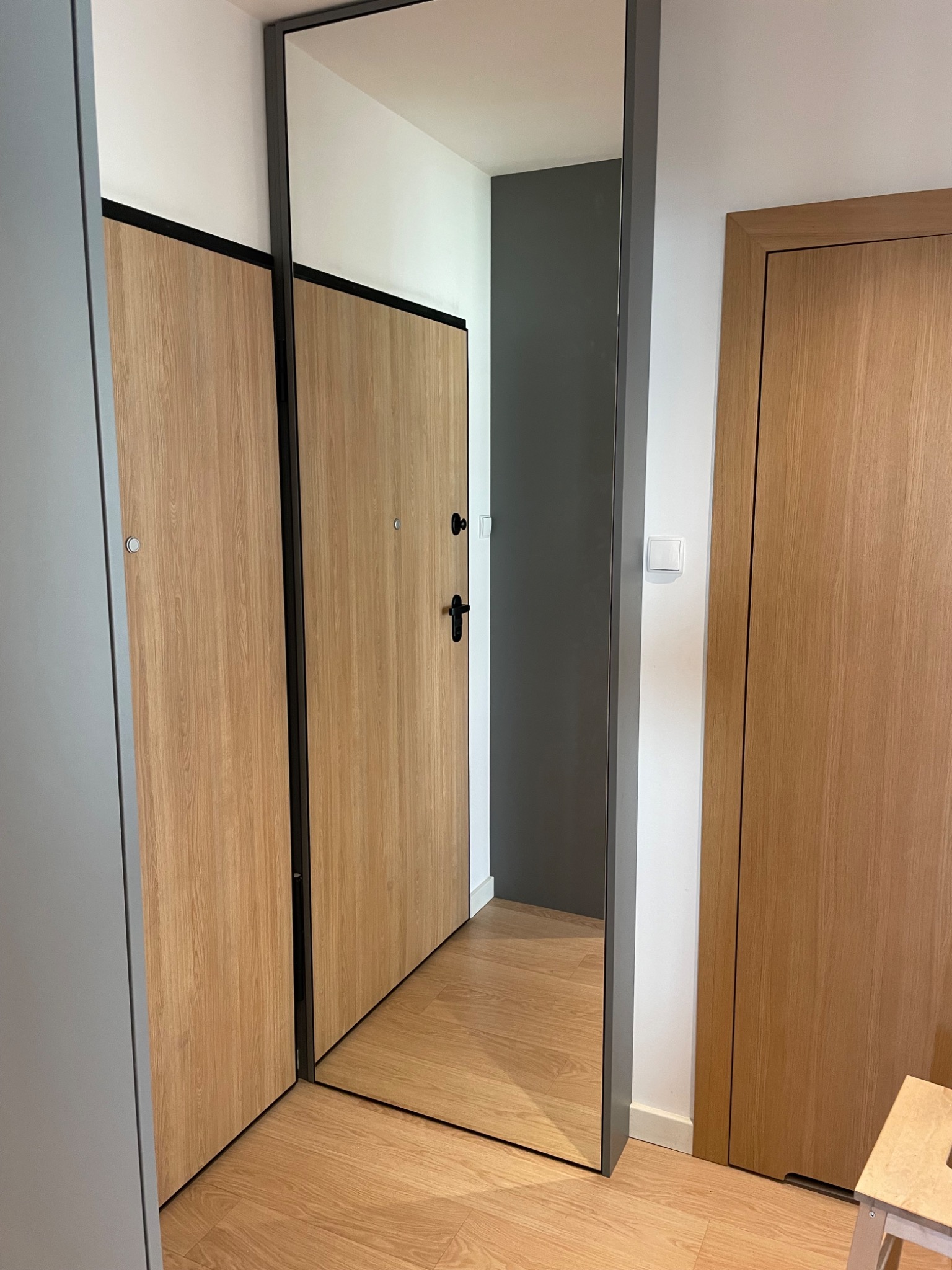 Wnętrze przedpokoju z dwoma drewnianymi drzwiami, jedno z nich odbija się w dużym lustrze w czarnej ramie, widoczny fragment podłogi i ściany w kolorze szarym.