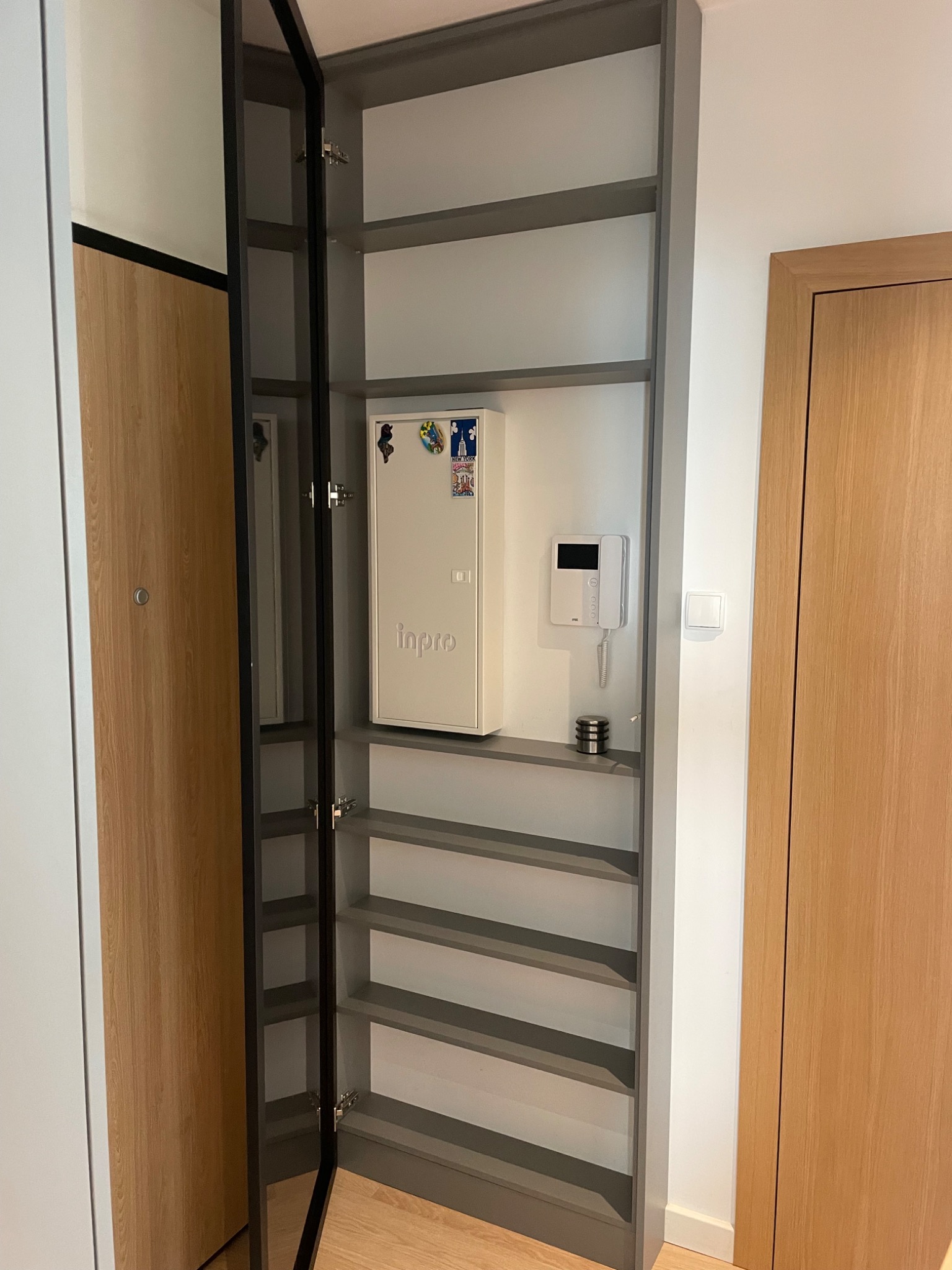 Wysoka, szara szafa wnękowa z otwartymi drzwiami, widoczne półki, na ścianie skrzynka elektryczna z magnesami i domofon, obok drzwi wejściowe.