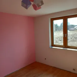 Pokój po odświeżeniu: Jedna ściana pomalowana na różowo, drewnopodobna podłoga, drewniane okno z widokiem na pole.