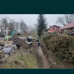 Wykopany rów z wodą, w którym stoi mężczyzna w gumowych butach, w tle wkopana rura, na górze koparka i usypana ziemia, w oddali dom.