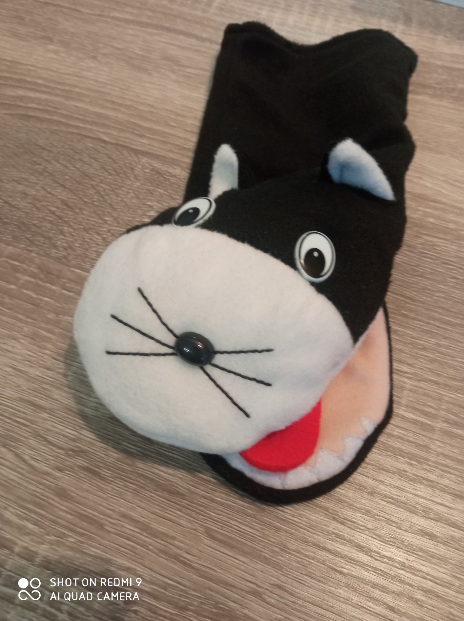 Czarno-biała pacynka w kształcie kota z wystającą czerwoną częścią imitującą język, leżąca na drewnianej powierzchni.
