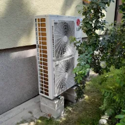 Zewnętrzna jednostka pompy ciepła LG Therma V, zamontowana na betonowych blokach przy ścianie domu z elewacją w kolorze beżowym, częściowo zasłonięta przez krzew z zielonymi liśćmi.