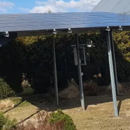 Konstrukcja wsporcza z panelami słonecznymi w ogrodzie, widoczne stalowe słupy nośne, trawnik, krzewy i dom w tle.