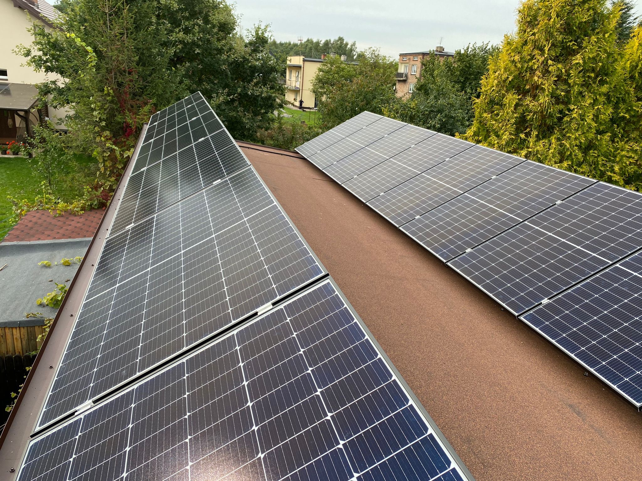 Panele słoneczne zamontowane na brązowym dachu dwuspadowym, widok z góry na tle zieleni drzew i budynków mieszkalnych.