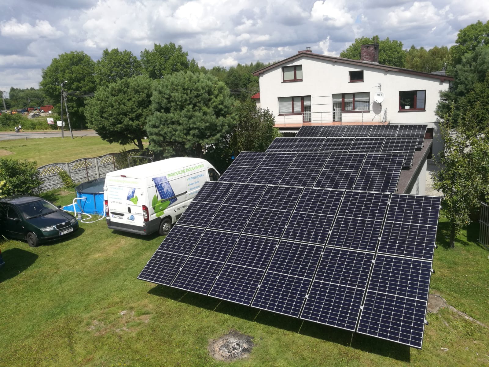 Panel słoneczny na dachu domu jednorodzinnego, obok zaparkowany samochód i bus z logo firmy, w tle drzewa i błękitny basen.