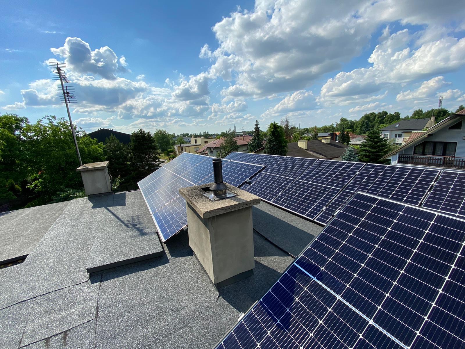 Panele słoneczne na dachu domu jednorodzinnego, widok z góry, błękitne niebo z chmurami w tle, widoczna antena telewizyjna i komin.