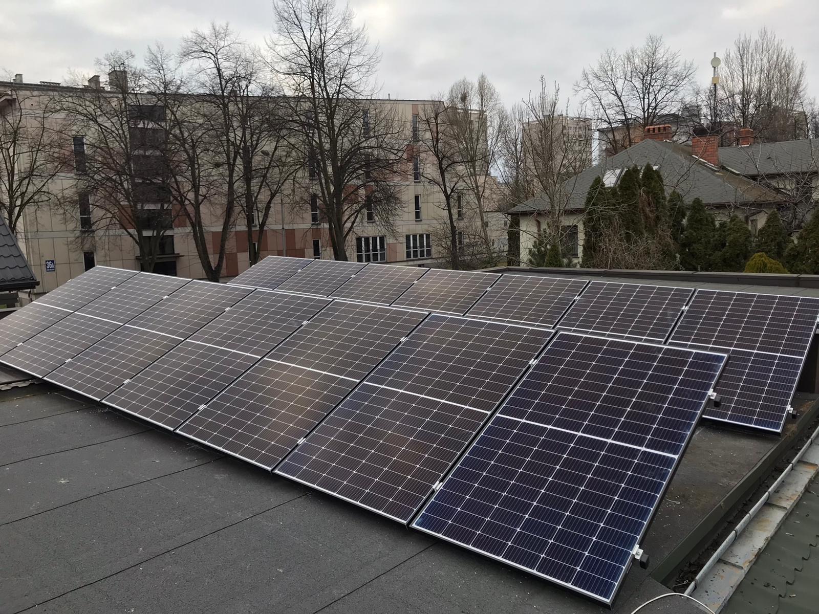 Panele słoneczne zamontowane na płaskim dachu budynku, widoczne budynki mieszkalne w tle, pochmurne niebo.