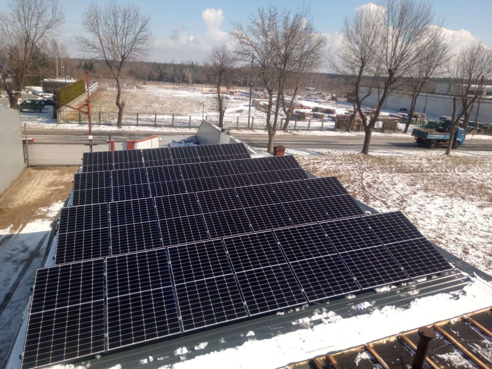 Instalacja fotowoltaiczna na dachu budynku przemysłowego pokrytym śniegiem, widok z góry, w tle drzewa i droga z przejeżdżającą ciężarówką.
